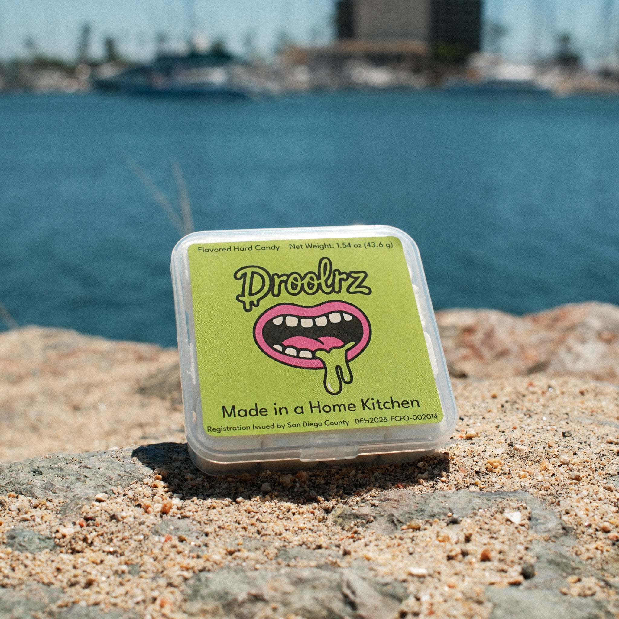 A pack of Droolrz candy sitting on a sandy rock with the ocean in the background